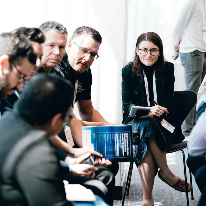 A group of people sit in a circle having a discussion; one woman with glasses smiles while holding a notebook.