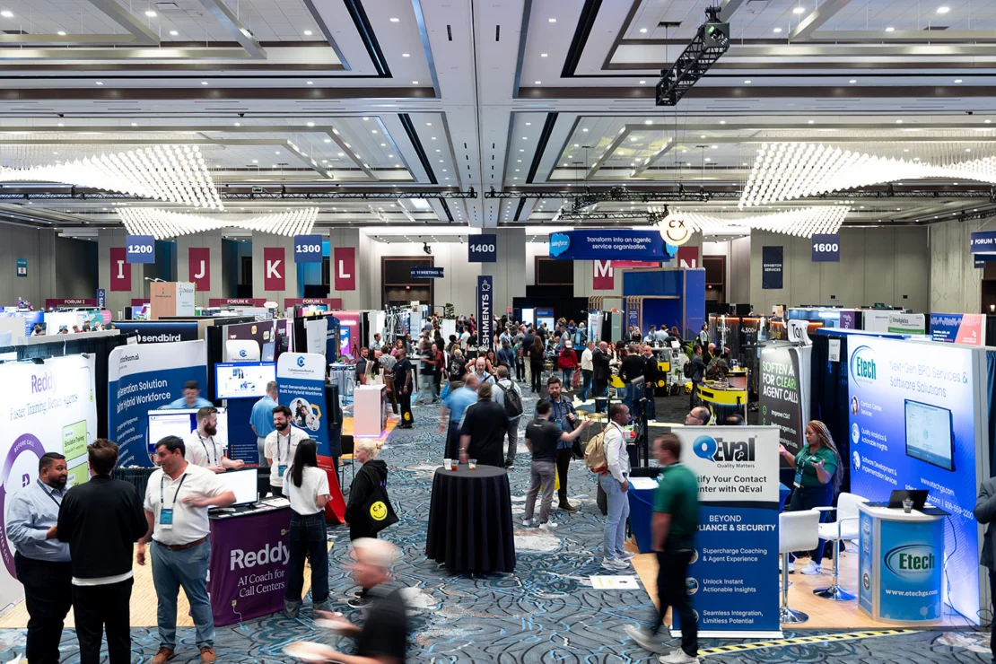 A busy convention center exhibits hall with people walking, networking, and visiting various company booths under bright lights.