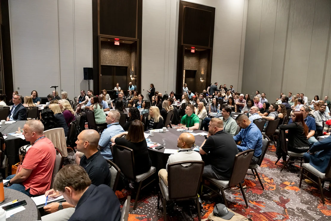 Large group of people seated at round tables in a conference room, attentively listening to a presentation or speaker.