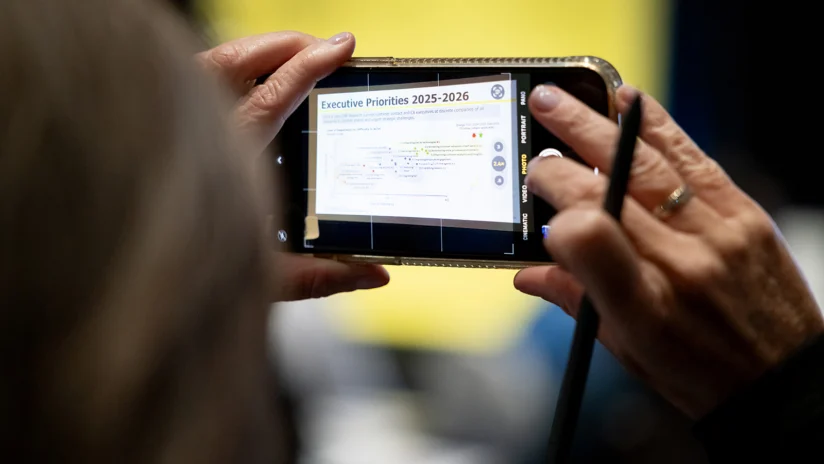 A person holds a smartphone and stylus, taking a photo of a screen displaying "Executive Priorities 2025–2026" and a list of bullet points.