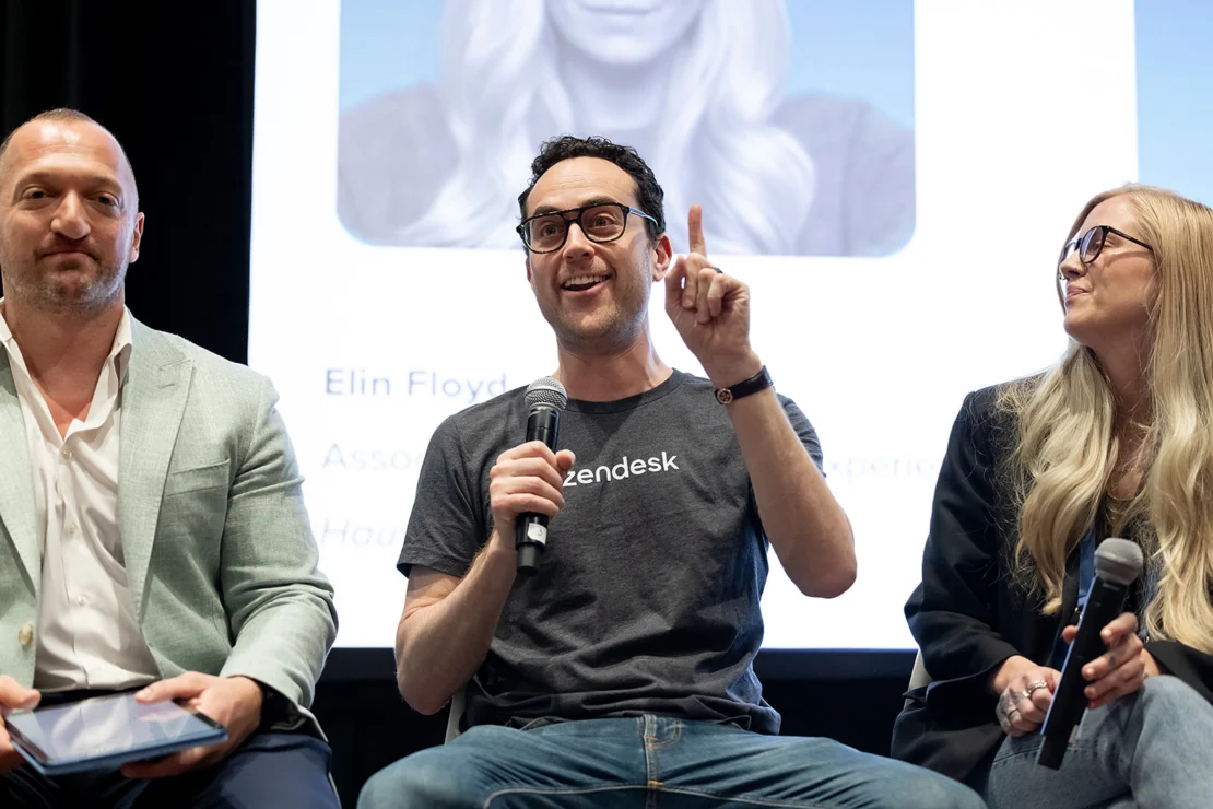 Three people sit on a panel; the man in the center, holding a microphone and gesturing upward, wears a Zendesk shirt. The others listen attentively. A blurred projection is in the background.