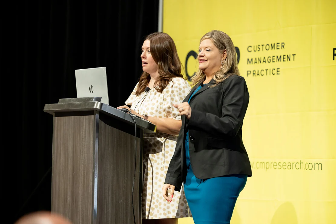 Two women stand on stage, one speaking at a podium with a laptop, in front of a yellow backdrop with "Customer Management Practice" text.