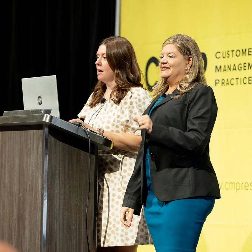 Two women stand on stage, one speaking at a podium with a laptop, in front of a yellow backdrop with "Customer Management Practice" text.