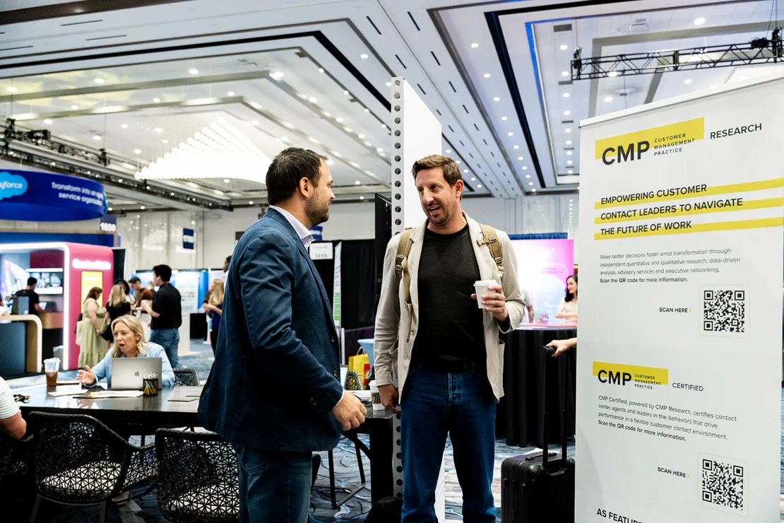 Two men stand and talk near a CMP research banner at a conference; other attendees work or converse at tables in the background.
