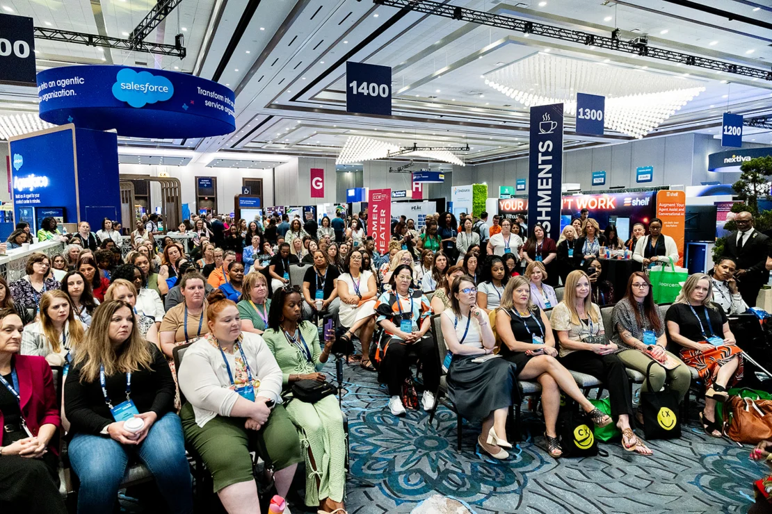A large group of people seated and listening at a conference event in a spacious, well-lit venue with various banners and booths in the background.