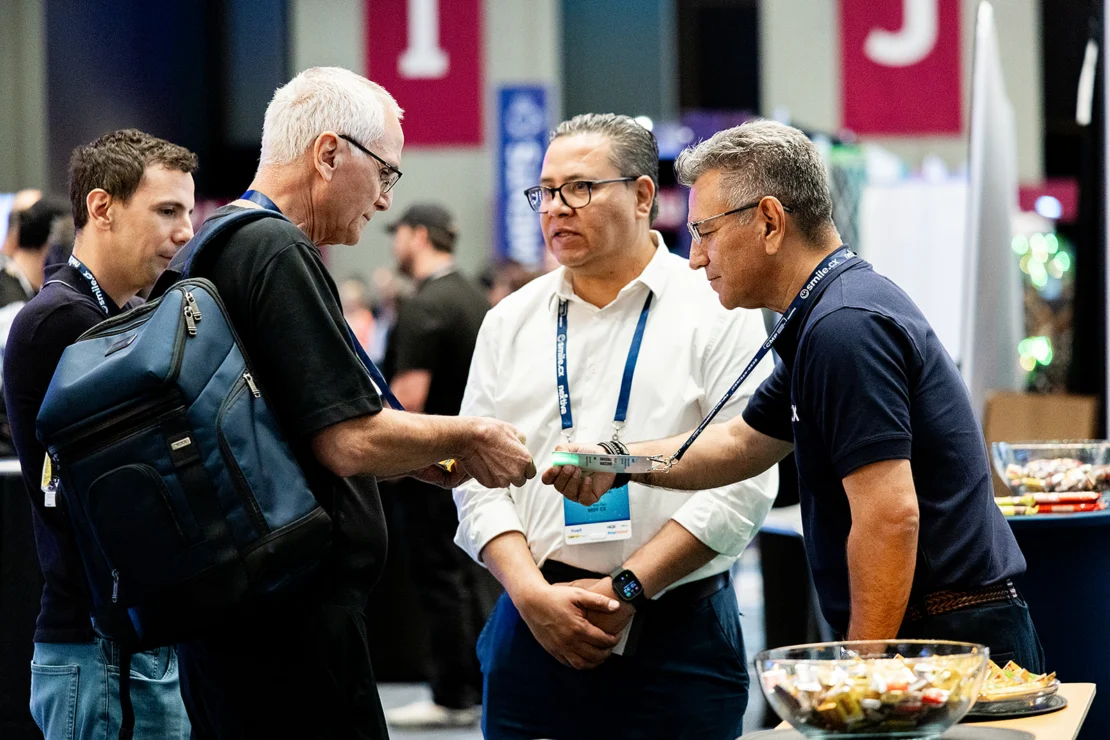 Four men, wearing conference badges, stand together conversing indoors; one man hands an item to another, with bowls of snacks visible on the table nearby.