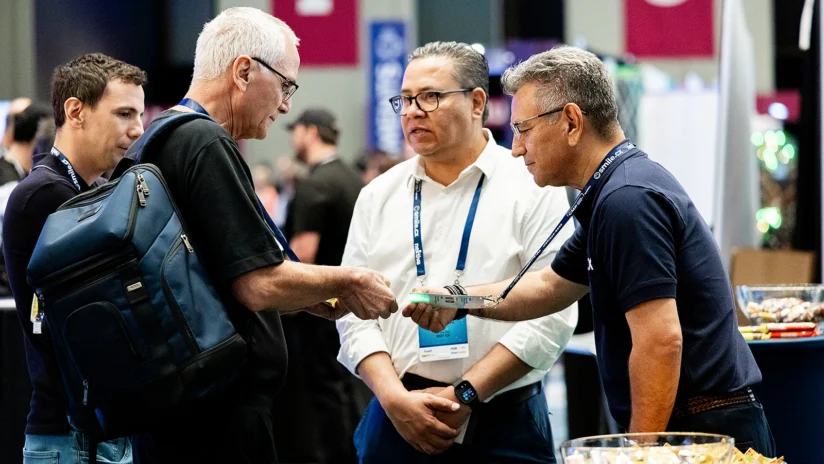 Four men, wearing conference badges, stand together conversing indoors; one man hands an item to another, with bowls of snacks visible on the table nearby.