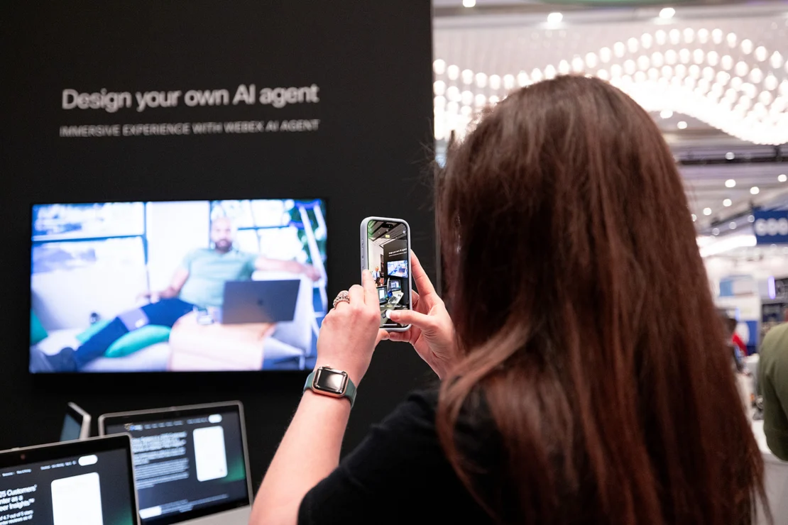 A person takes a photo with a smartphone at an exhibit booth displaying the text "Design your own AI agent" and a screen showing a man sitting with a laptop.