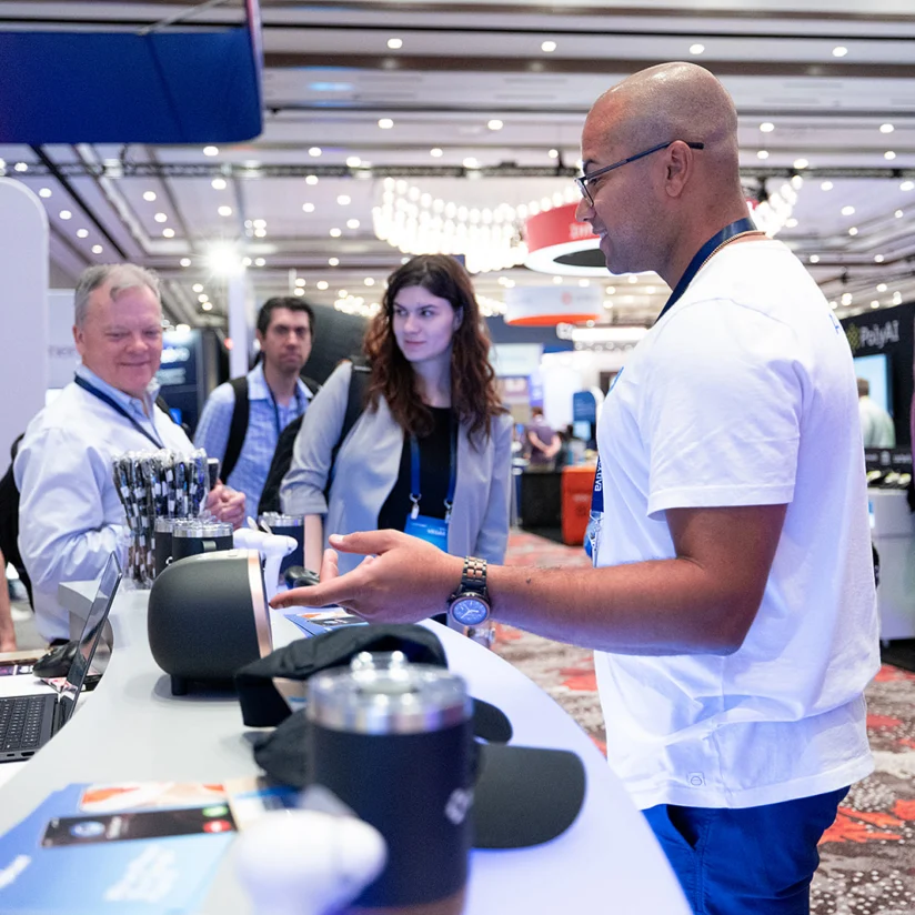 A man in a white shirt speaks to several people at a booth during a conference or trade show in a large, well-lit room.