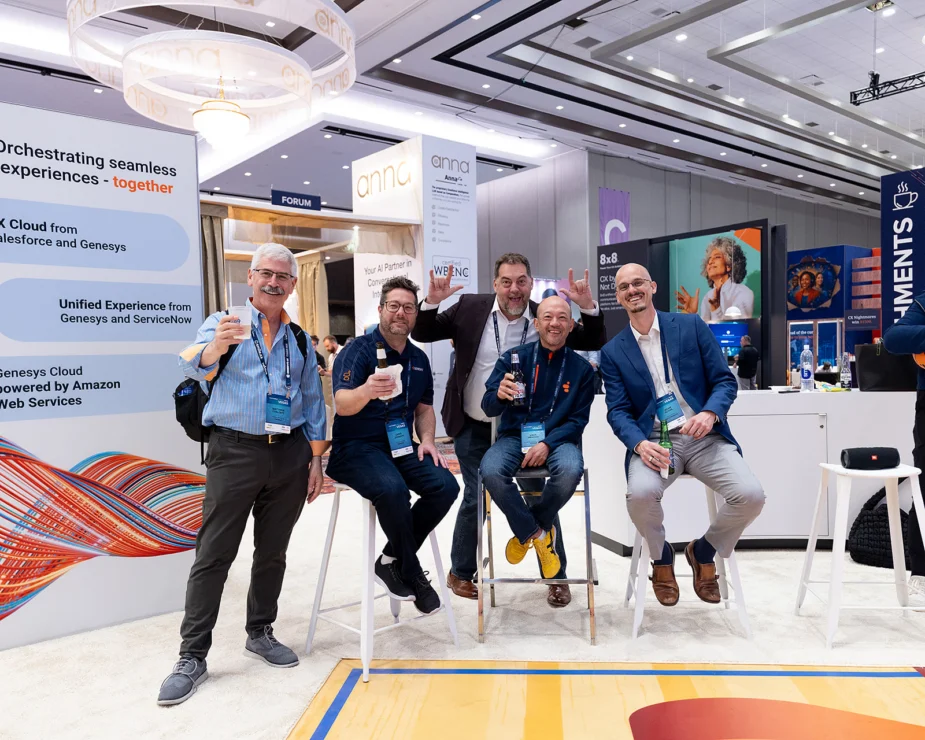 Five men pose together and smile while holding drinks in a brightly lit exhibition hall with tech company booths and banners in the background.