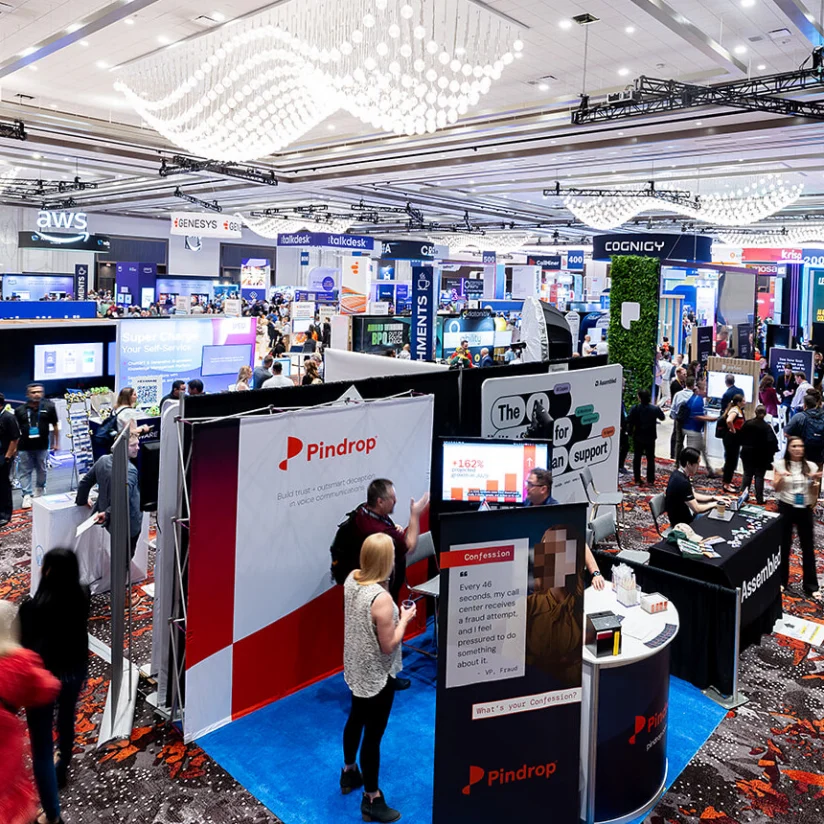 Crowded exhibition hall with multiple company booths, attendees walking, and various displays under bright ceiling lights at a technology conference.