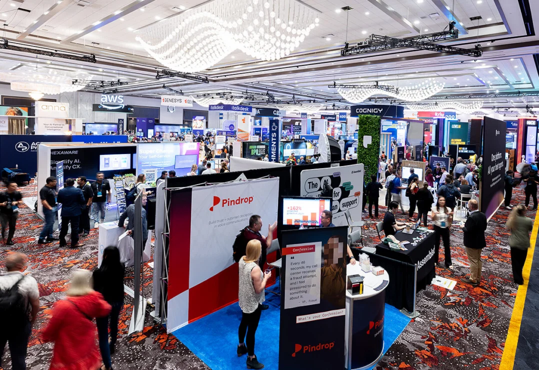 Large technology conference expo hall with multiple company booths, displays, and attendees walking and interacting in a brightly lit, spacious venue.