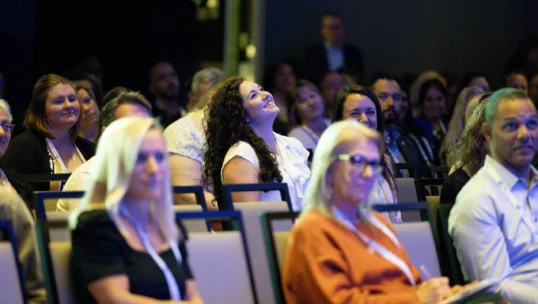 Audience members sit in rows of chairs at an indoor event, with several people looking towards the stage or screen.
