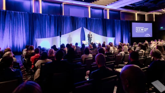 A speaker stands on stage addressing an audience in a conference room with a large screen displaying the CCW logo.