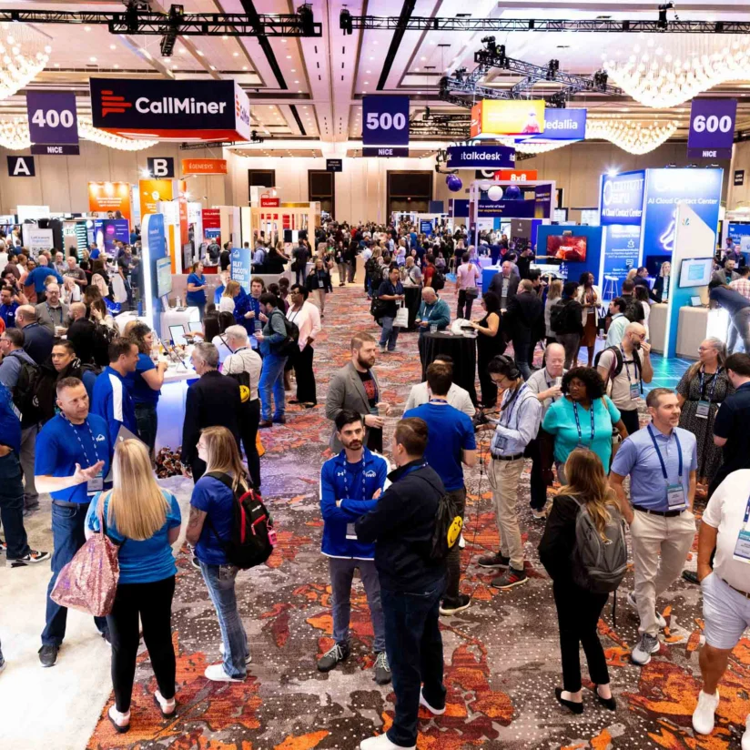 Large crowd of people networking and visiting booths at a busy business conference or trade show in a spacious, well-lit exhibition hall.