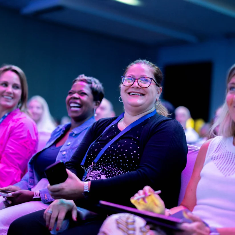 A group of four women sit in an audience, smiling and laughing, with conference badges around their necks in a brightly lit room.