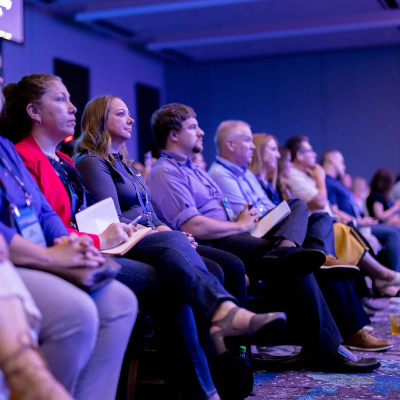 A group of people sit closely together in rows, attentively watching a presentation or event in a dimly lit conference room.