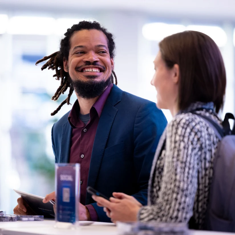 A man in a blue blazer and a woman with a backpack are talking at a counter, both holding electronic devices.