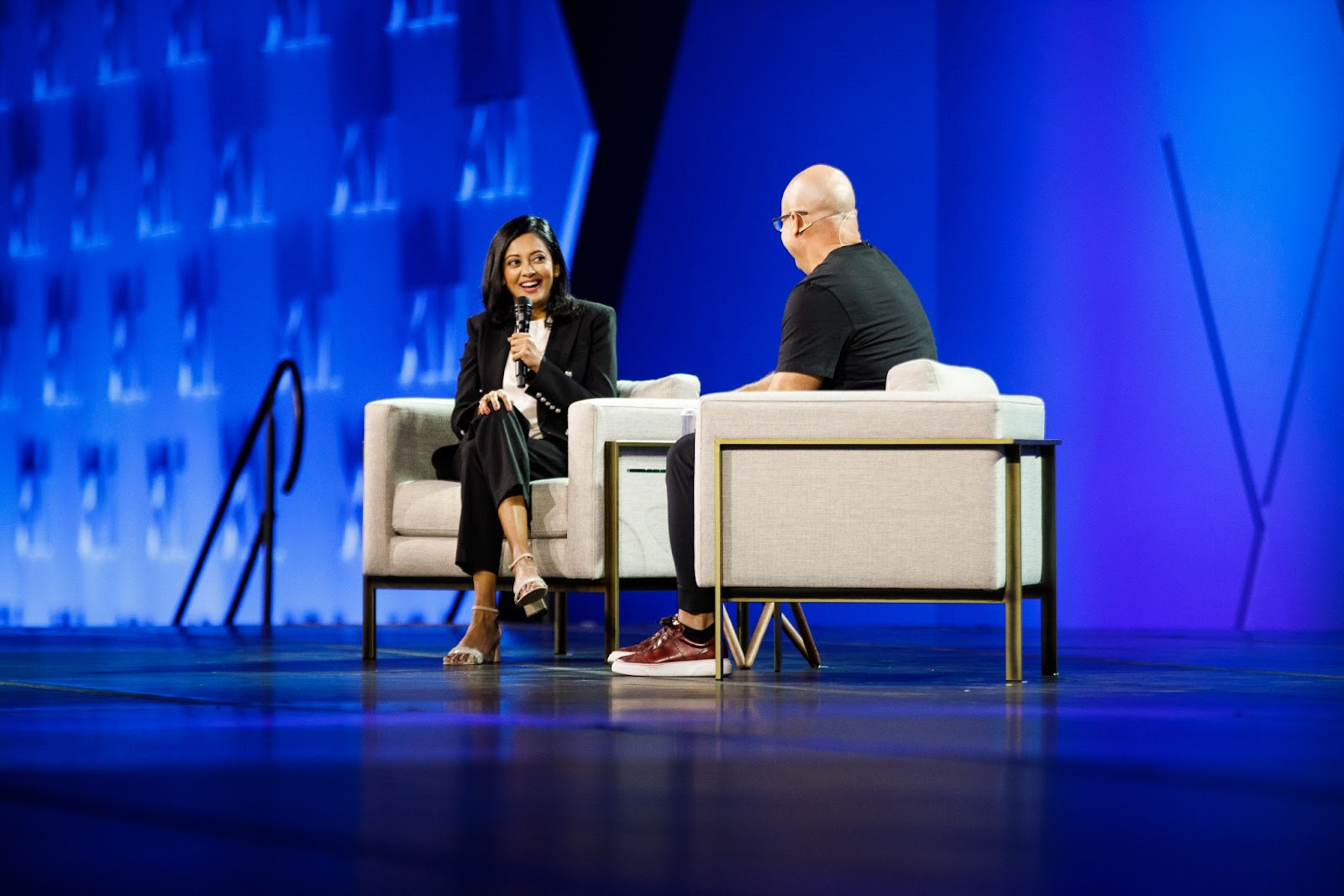 Two people sit on stage in armchairs having a conversation, with one holding a microphone, against a blue-lit background.