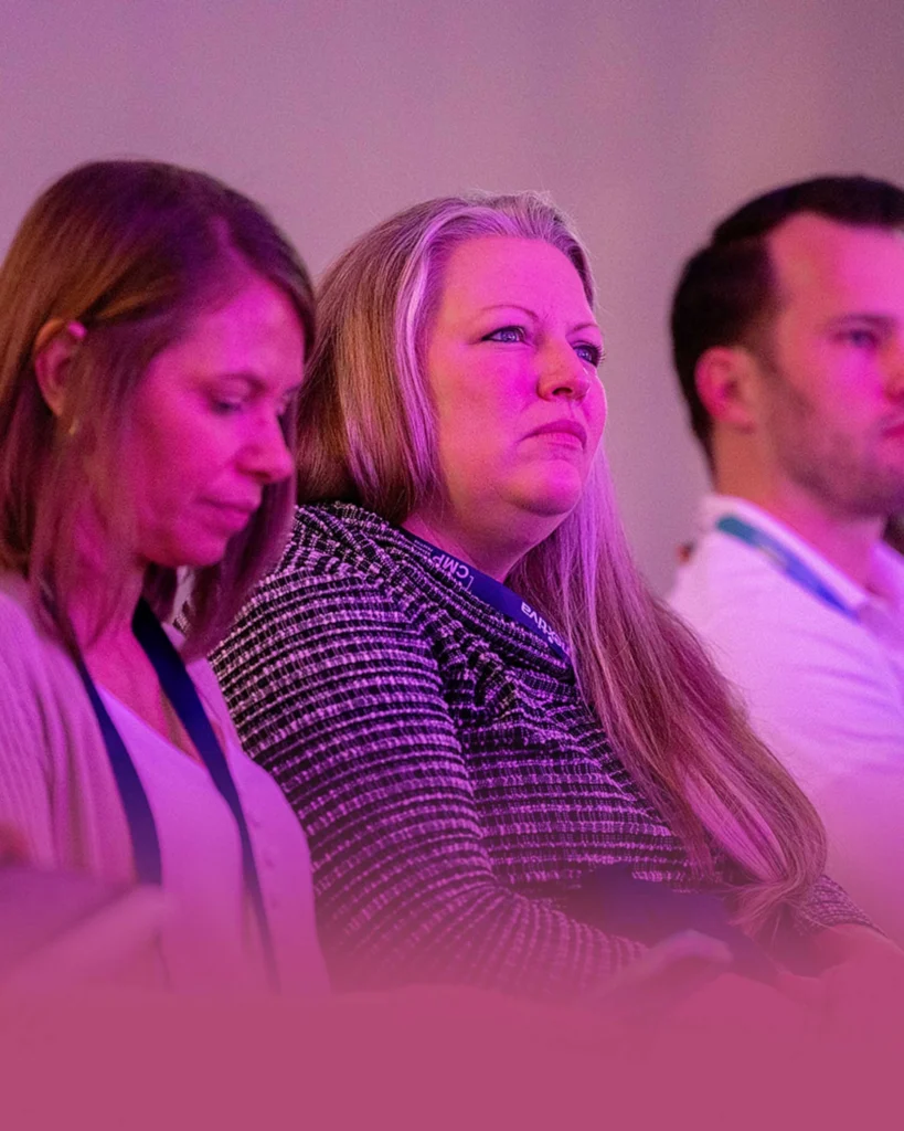 Three people sit in a row at an indoor event, attentively facing forward; the image has a purple-pink lighting effect.
