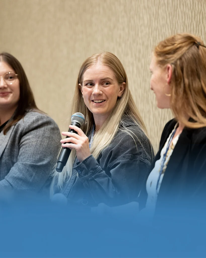 Three women sit next to each other at a panel discussion; the woman in the center holds a microphone and smiles while speaking to the woman on her right.