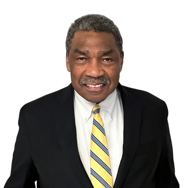 A man wearing a black suit, white shirt, and yellow striped tie stands against a plain white background, looking at the camera and smiling.