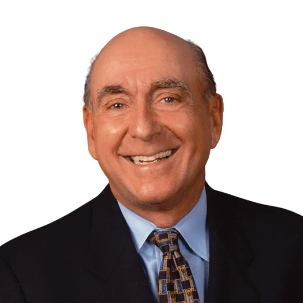 A smiling bald man wearing a dark suit, light blue shirt, and patterned tie against a white background.