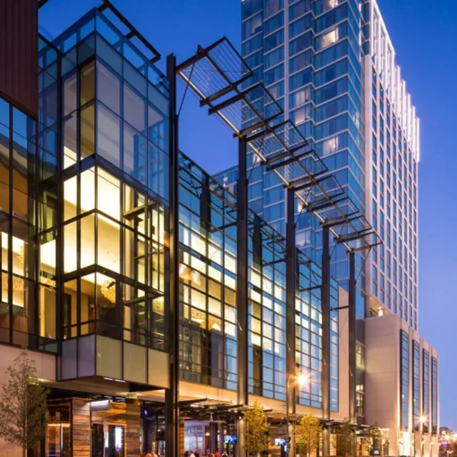 Modern glass-fronted high-rise building with illuminated lower levels and a geometric metal canopy, pictured at dusk.