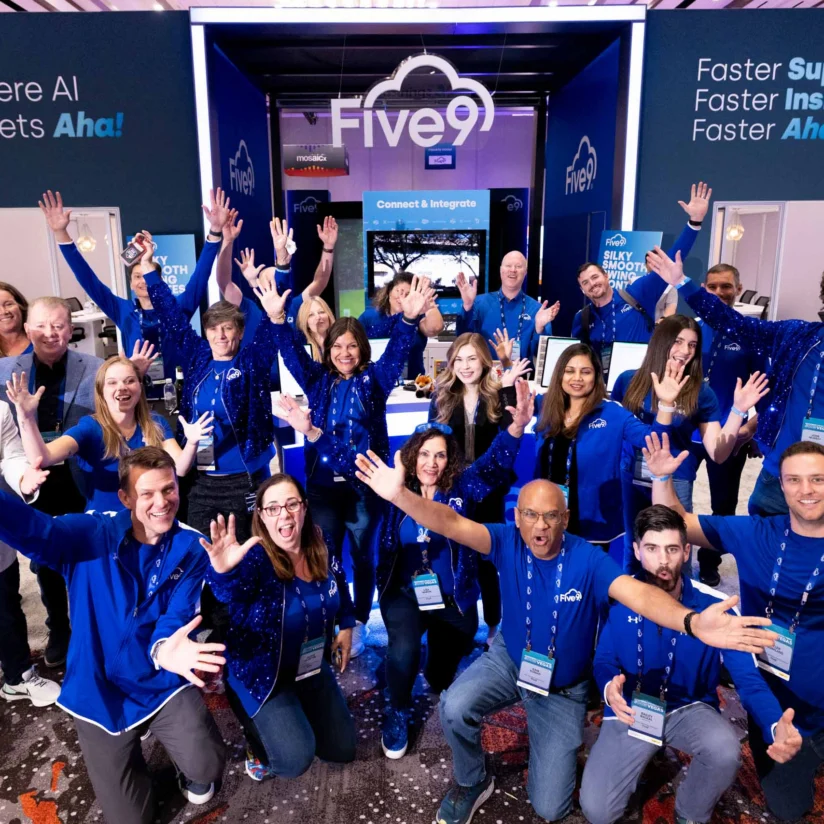 A group of people wearing blue shirts pose with raised hands in front of a Five9 booth at a conference or trade show.