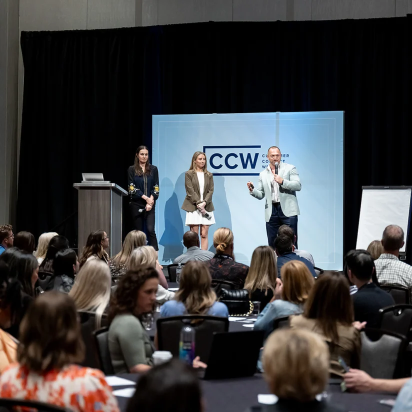 Three presenters stand on stage in front of a seated audience at a conference, with a CCW sign and a blank flip chart in the background.