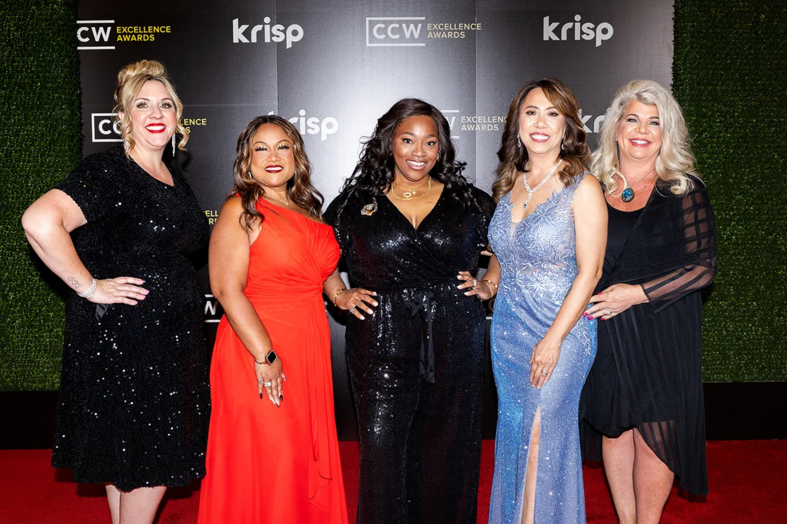 Five women stand side by side on a red carpet, dressed in formal evening gowns, posing for a photo at an awards event with branded banners in the background.