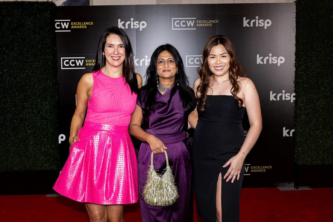 Three women in formal dresses pose and smile on a red carpet in front of a CCW Excellence Awards and Krisp event backdrop.