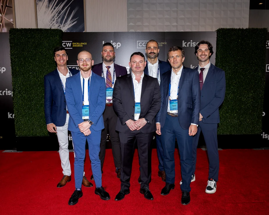 Seven men in business attire stand on a red carpet in front of a green and black event backdrop displaying logos and signs.