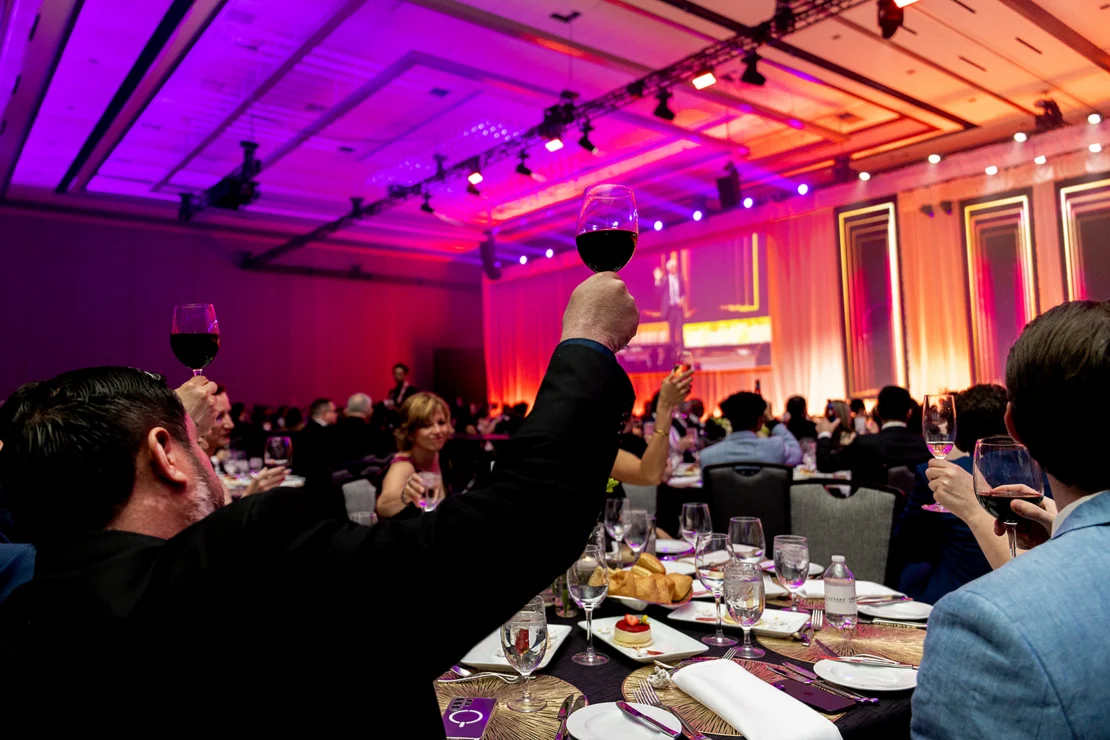People raise glasses of red wine in a toast at a formal banquet with decorated tables, stage lighting, and a large crowd in a spacious event hall.