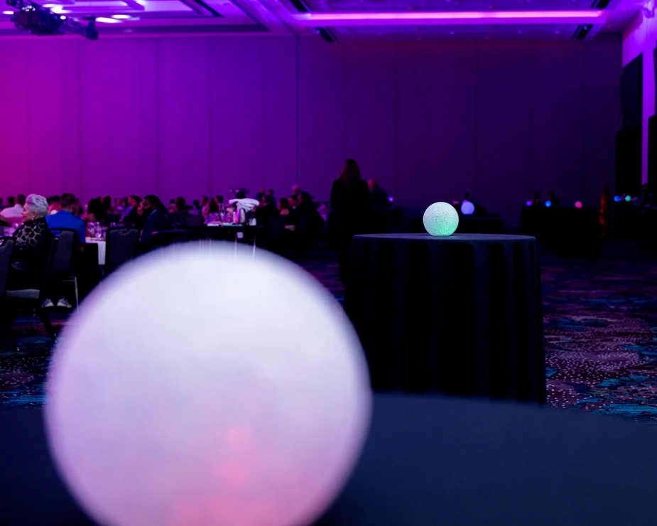 A dimly lit banquet hall with round tables, people seated in the background, and glowing decorative spheres on tables in the foreground and middle ground.