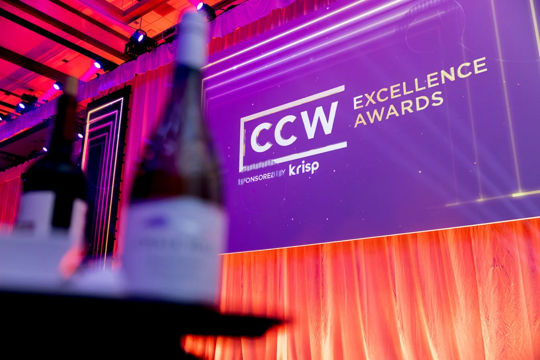 Large screen displaying "CCW Excellence Awards" on a stage with pink-orange curtains and spotlights; wine bottles are blurred in the foreground.
