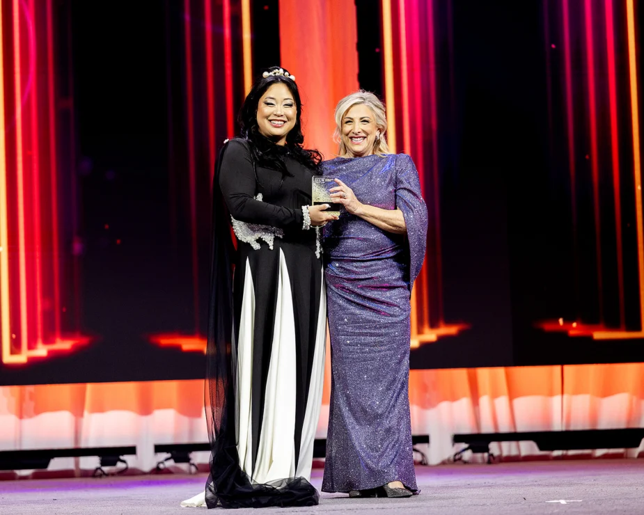 Two women stand on stage, smiling and holding an award together, with a colorful, illuminated backdrop behind them.