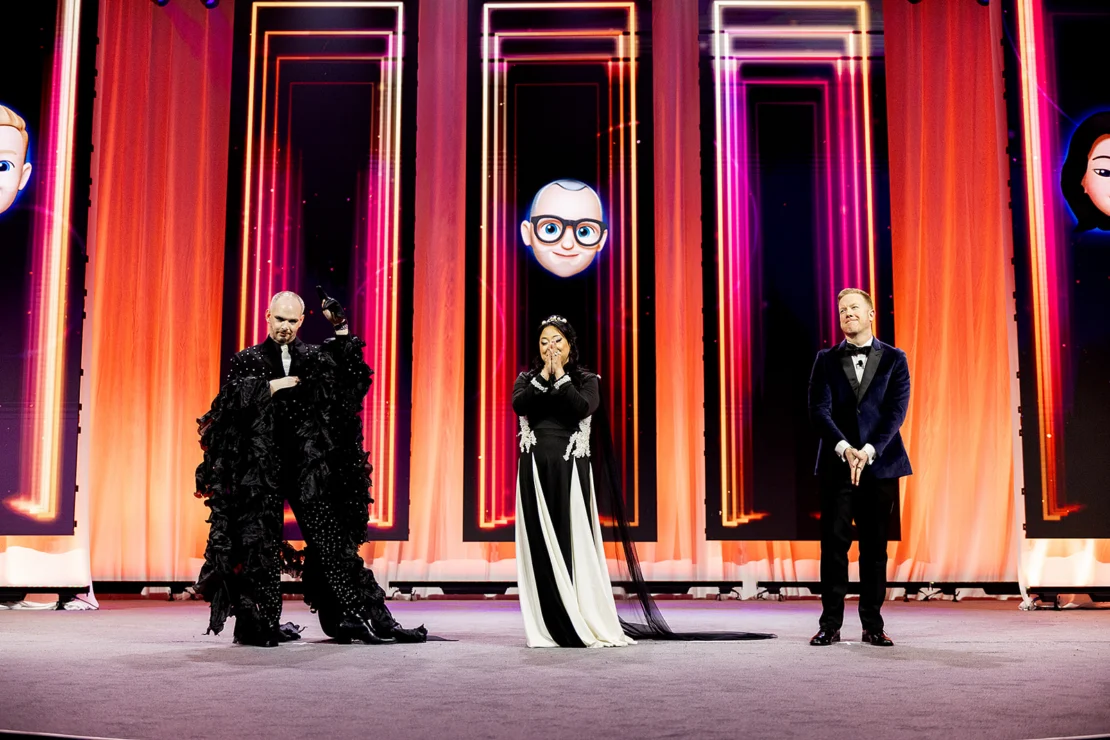 Three people stand on stage in formal attire with animated avatar faces displayed on large screens behind them, against a backdrop of vertical illuminated panels.