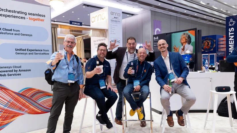 Five men pose and smile while sitting and standing at a trade show booth, holding drinks. Conference signage, banners, and booths are visible in the background.