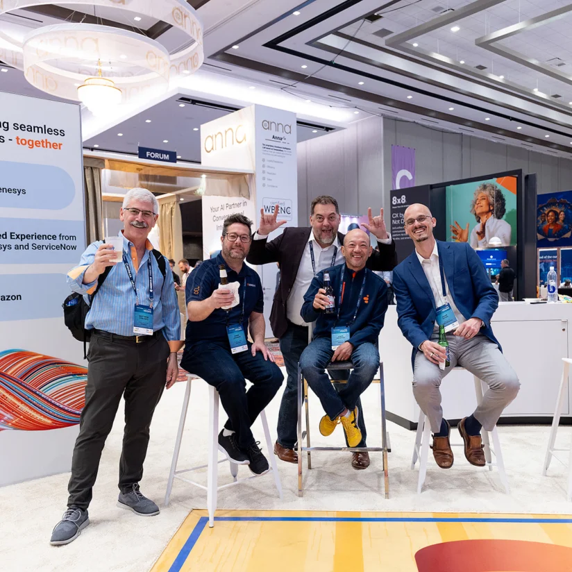 Five men pose and smile while sitting and standing at a trade show booth, holding drinks. Conference signage, banners, and booths are visible in the background.