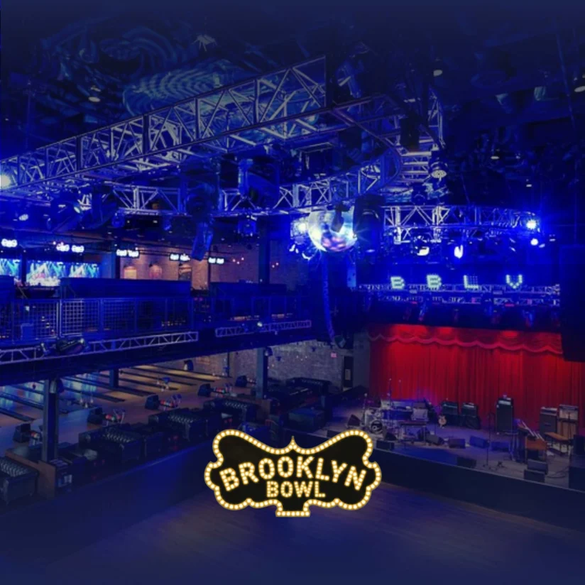 A view of the Brooklyn Bowl venue with a stage, bowling lanes, and blue lighting, featuring the Brooklyn Bowl logo in the foreground.