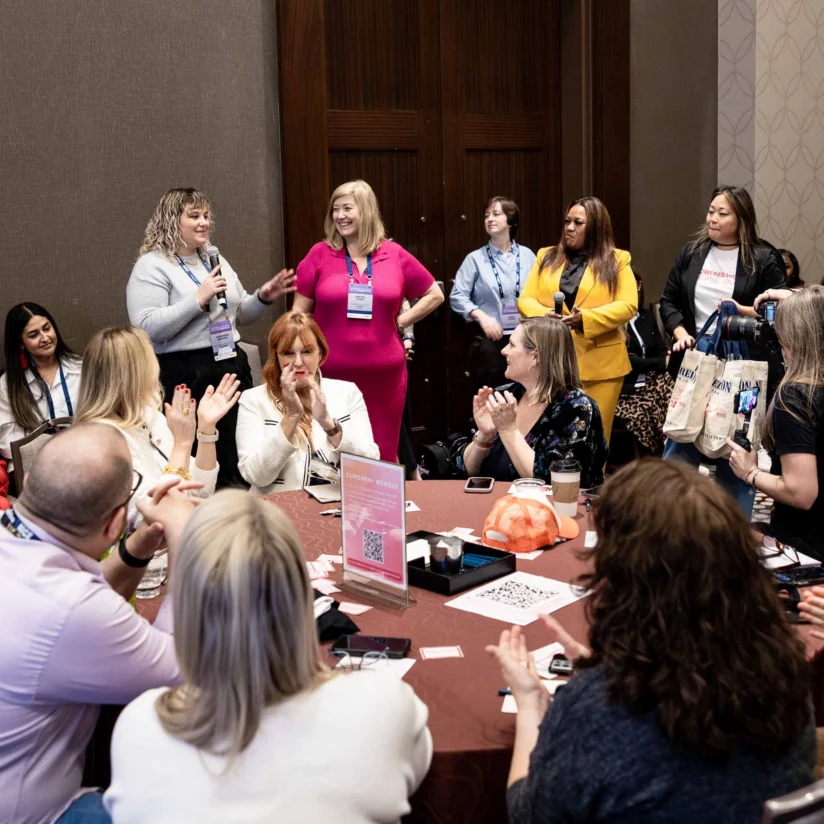 A group of people seated around tables in a conference room listen and applaud as two women stand and speak at the front of the room.