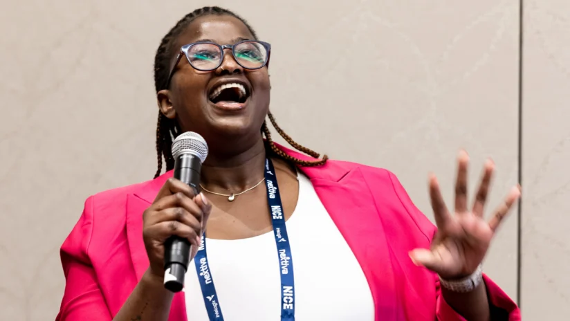 A person wearing a pink blazer and glasses speaks into a microphone, smiling and gesturing with one hand during a presentation.