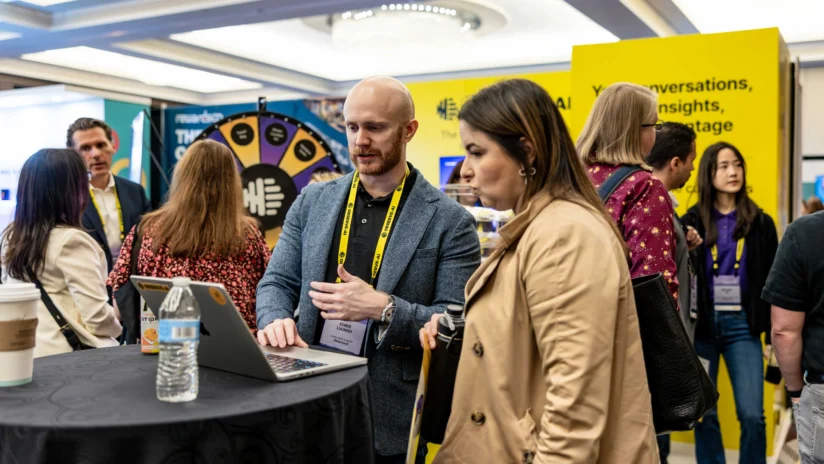 People at a conference booth interact with a laptop; bright yellow banners and a prize wheel are visible in the background.