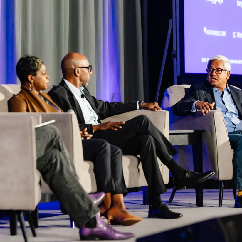 Four people sit in armchairs on a stage engaged in a panel discussion, with a blue curtain and presentation screen in the background.