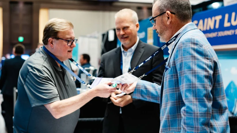 Three men in business attire stand in a conference setting, exchanging business cards and conversing near a booth.