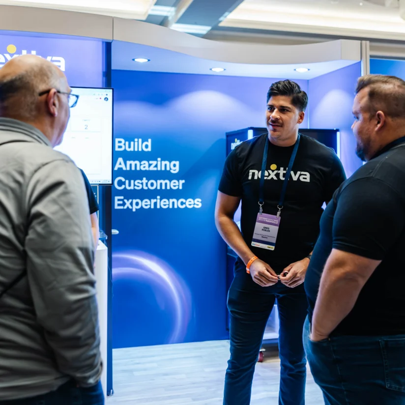Three men stand and talk at a trade show booth with a blue Nextiva display that reads "Build Amazing Customer Experiences.
