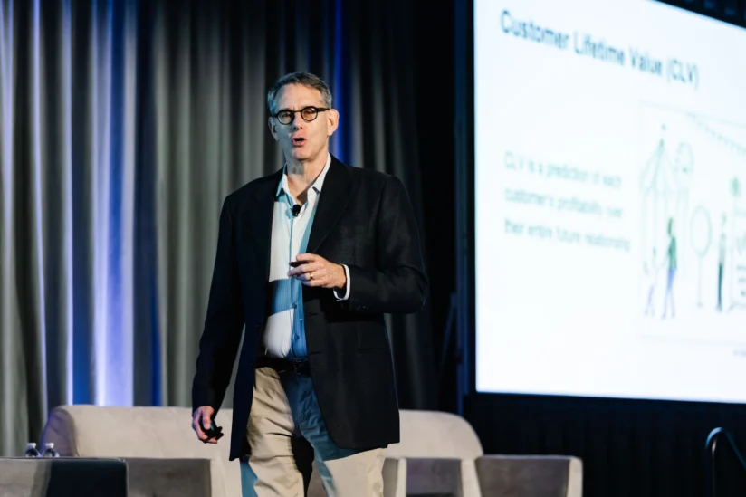 A man in a dark blazer and glasses speaks on stage near a projection screen displaying information about Customer Lifetime Value (CLV).