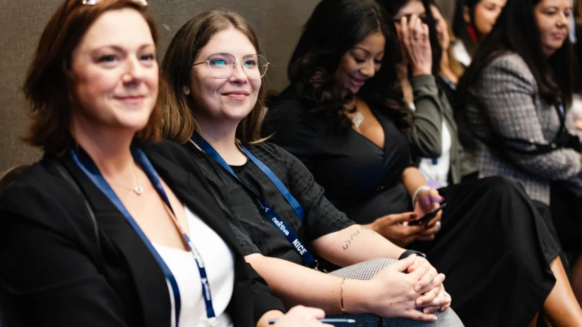A group of women sit in a row at an indoor event, some smiling at the camera while others look at their phones or talk.