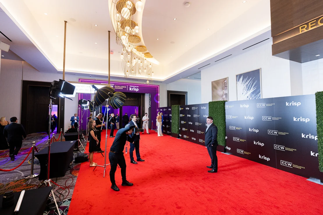 A man poses for photos on a red carpet at an indoor event, with photographers taking pictures and branded banners in the background.